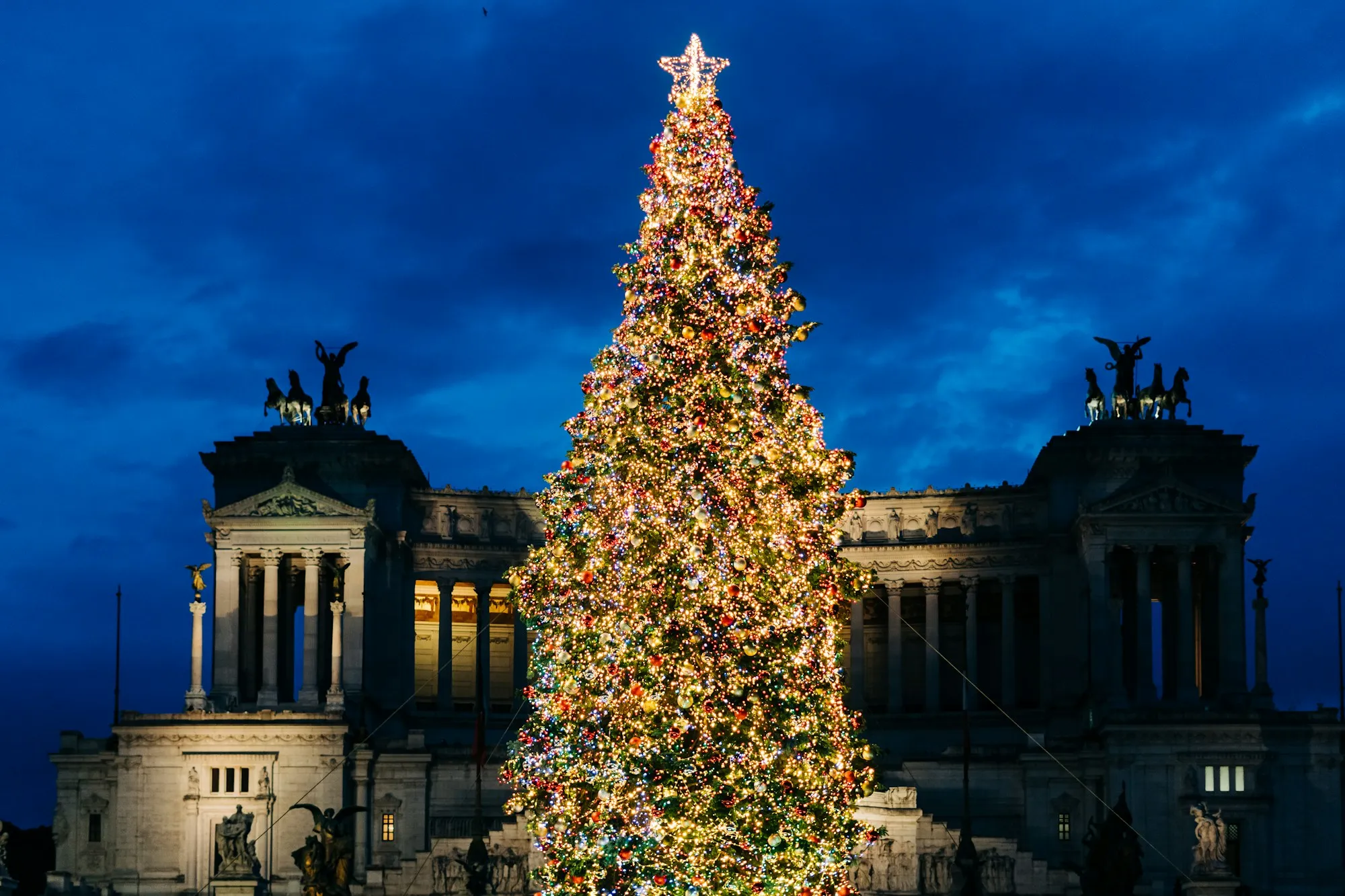 A tall Christmas tree lit at dusk in Piazza Venezia, with the white colonnade of the Vittoriano behind it